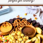 A round wooden board filled with autumn-themed snacks, including apple slices, pretzels, pecans, candy corn, popcorn, and chocolate cookies. Small bowls of caramel and peanut butter dips are placed on the board. Discover this Autumn Snack Board Recipe—perfect for sharing!.