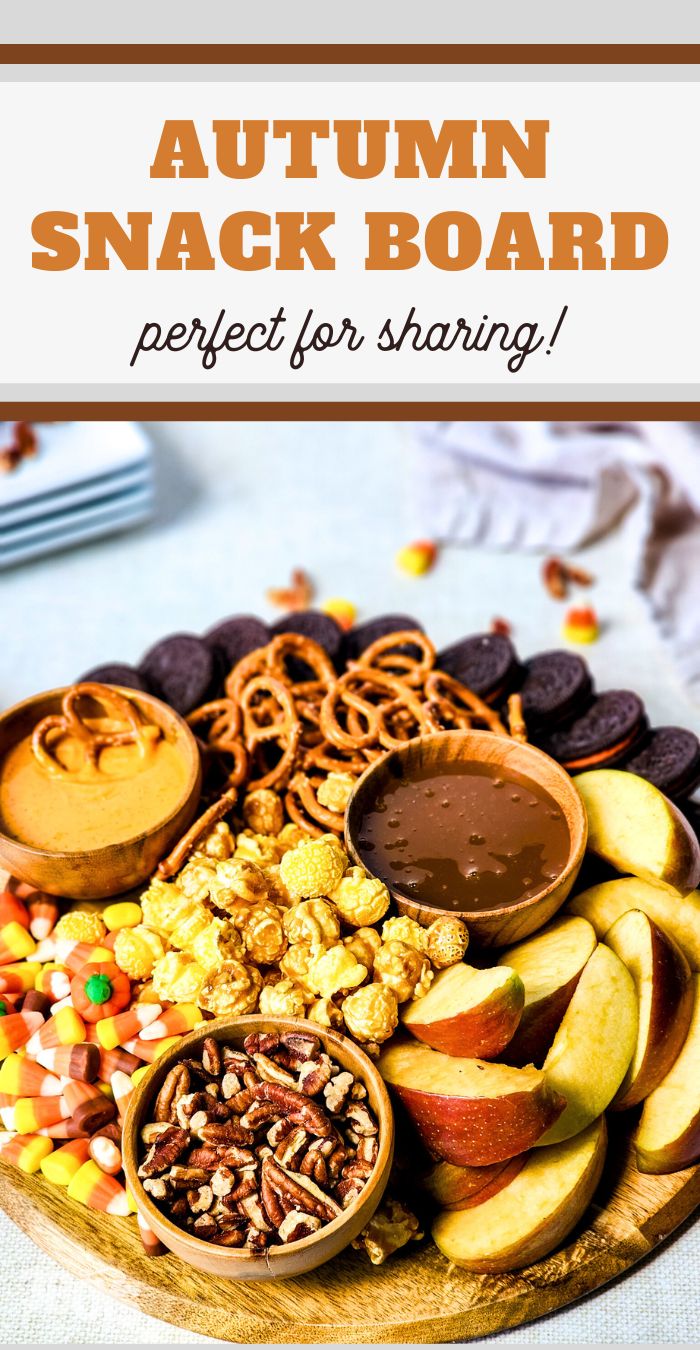 A round wooden board filled with autumn-themed snacks, including apple slices, pretzels, pecans, candy corn, popcorn, and chocolate cookies. Small bowls of caramel and peanut butter dips are placed on the board. Discover this Autumn Snack Board Recipe—perfect for sharing!.