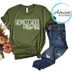 A green t-shirt with Homeschool Mama paired with distressed blue jeans. The setup, featuring autumn decorations like small pumpkins and leaves, complements the cozy learning space. A subtle nod to Free Solar System Printables can inspire creativity. The Amcee Designs logo sits in the top right corner.