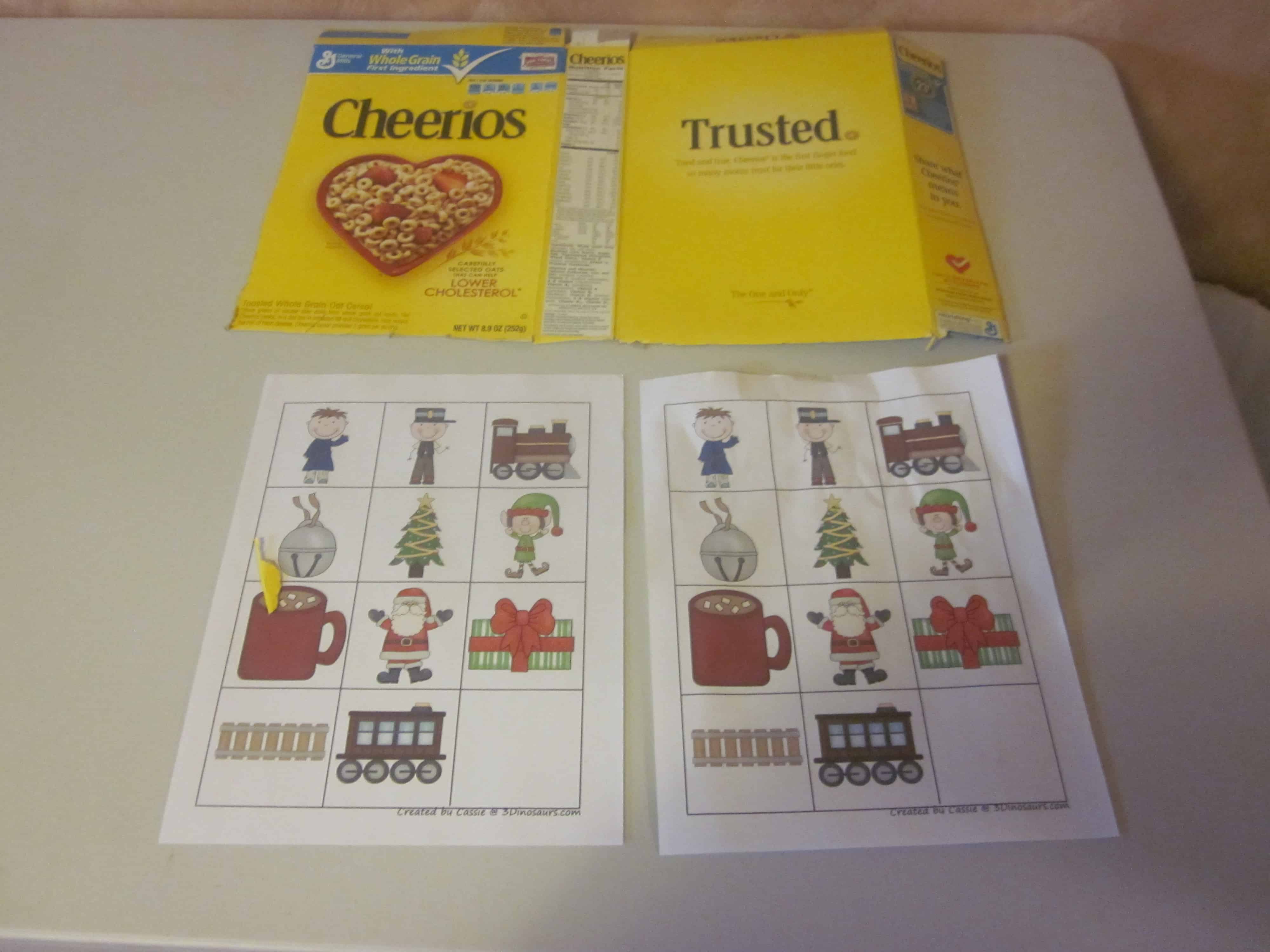 Two Cheerios cereal boxes, one open and one closed, are on a table. Below them are two sheets with a bingo-like game featuring images of a train, mug, Santa, gift, tree, and people.