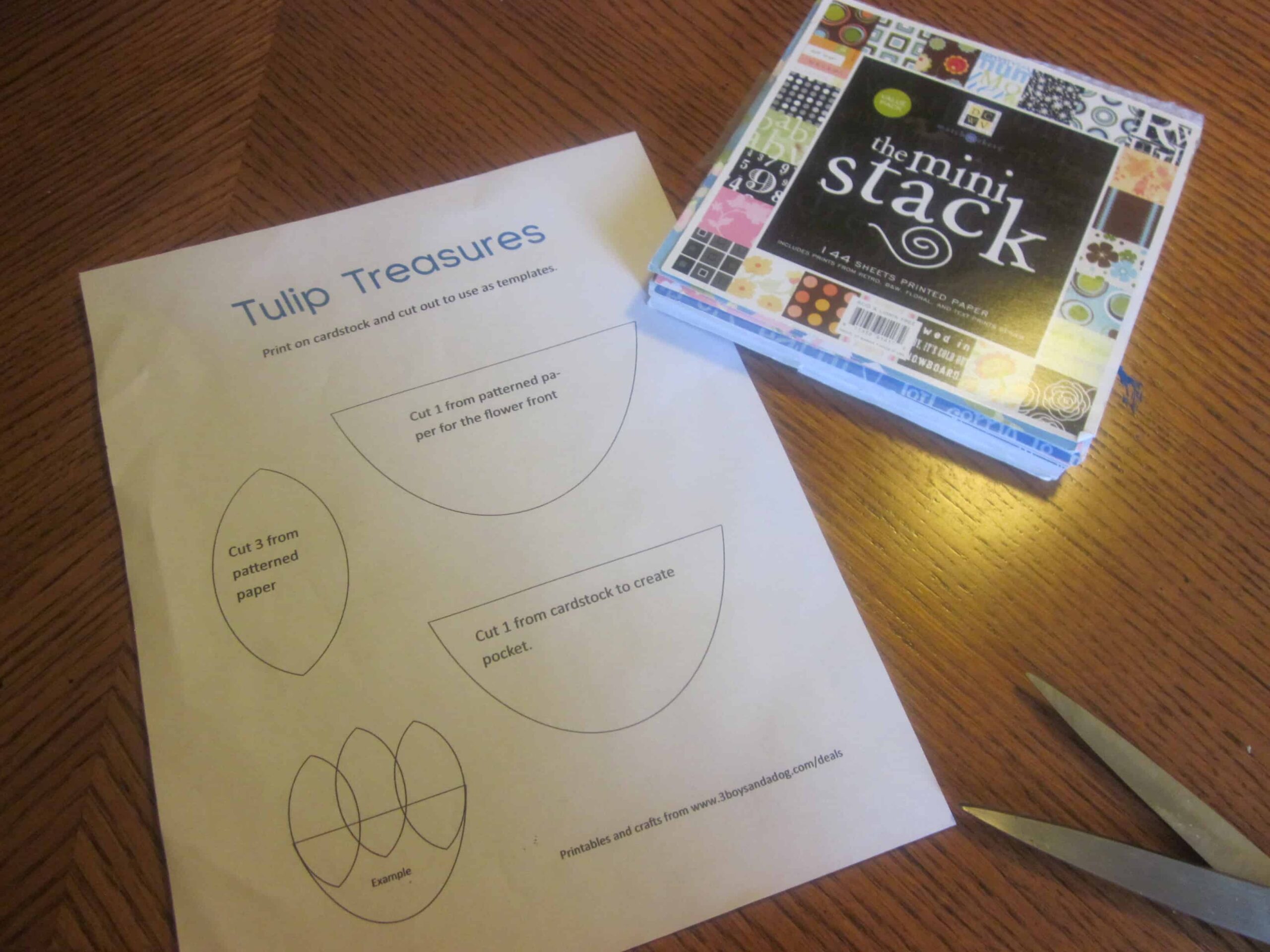 A sheet titled Tulip Treasures displays cutting templates for a DIY project on a wooden table. Perfect for Teacher Appreciation crafts, colorfully patterned paper pads labeled the mini stack and a pair of scissors sit ready to inspire creativity.