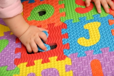 A childs hands are assembling colorful foam puzzle pieces with letters on them, aiding baby memory development. The textured pieces in vibrant colors like red, blue, green, purple, and yellow stimulate infant cognitive milestones as they play.