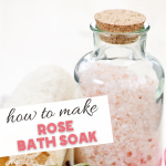 A glass jar filled with pink bath salts, sealed with a cork, showcases the Rose Bath Soak Recipe in elegant black and pink fonts. A hint of a rose and a loofah peek from the background, adding charm to this soothing creation.