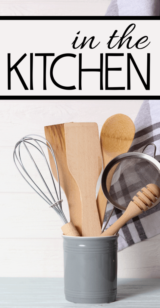 A collection of kitchen utensils in a gray container, including a whisk, wooden spoons, and a strainer, with the words in the KITCHEN above them on a white wooden background.
